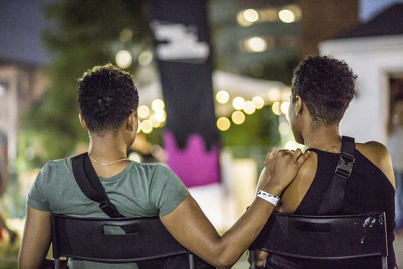 The backs of two people sitting in chairs, one with their hand on the shoulder of the other