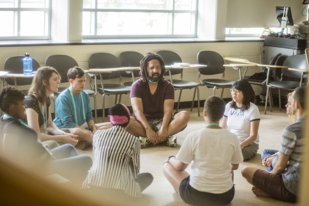 A group of nine people of various races sitting on the floor of a classroom. They have their eyes closed and appear to be singing or chanting.