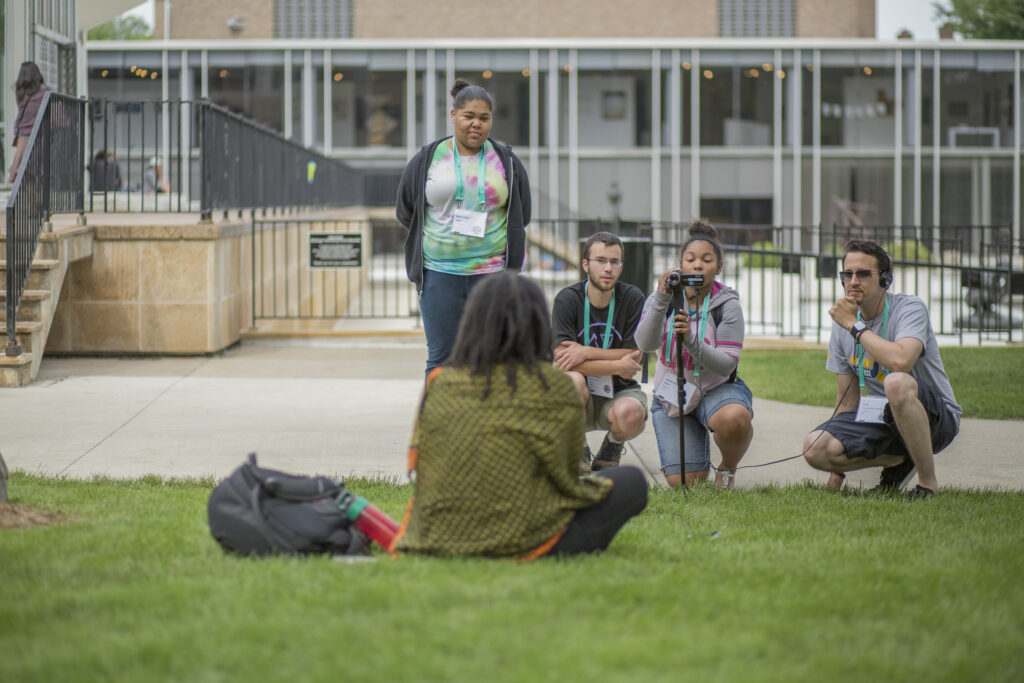 A group of four people filming another person outside on the grass.